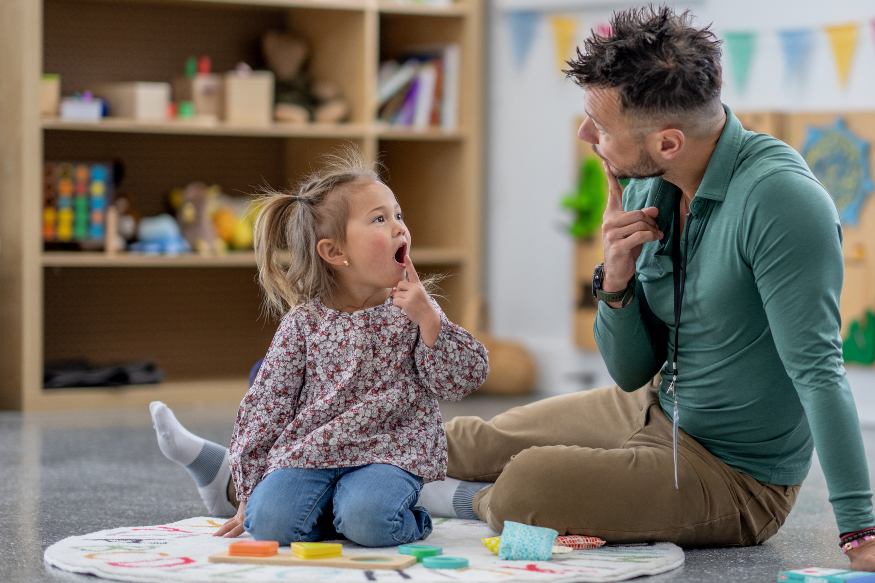 A male Speech therapist sits with a little girl on the floor as he works with her on phonetics. The therapist is making a sound and holding his finger to his mouth for the little girl to mimic.