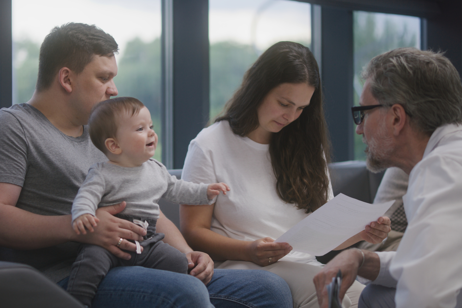 Family with little child sit on couch in clinic lobby area after doctors appointment. Doctor speaks about medical test results, gives papers.