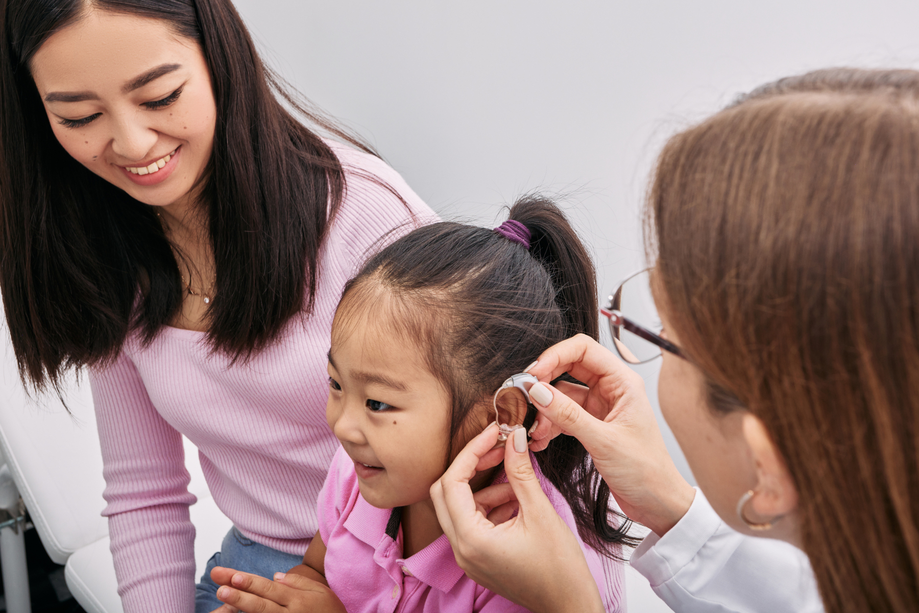 Audiologist fits hearing aid on Asian little girl's ear while visit in hearing clinic with mother. Hearing solutions for children