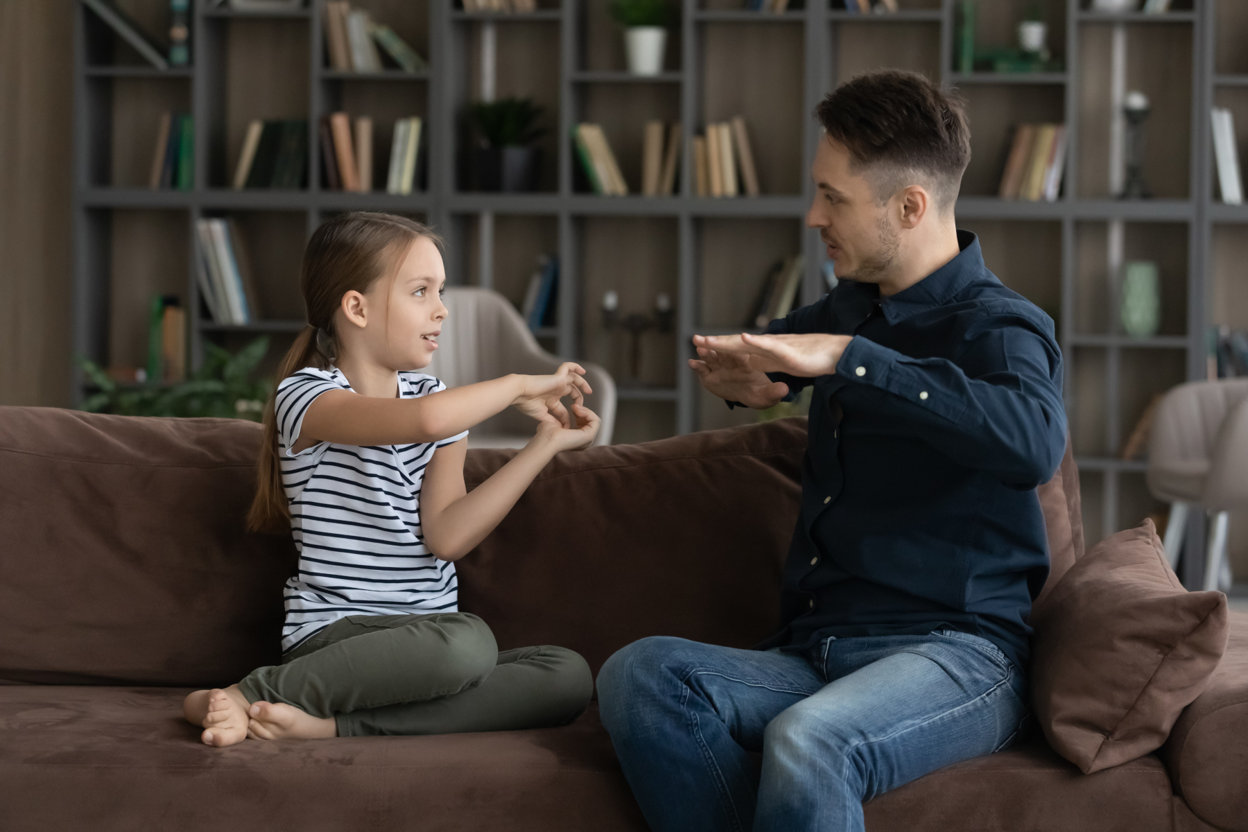 Little deaf girl and her father sit on couch speaking using sign language.